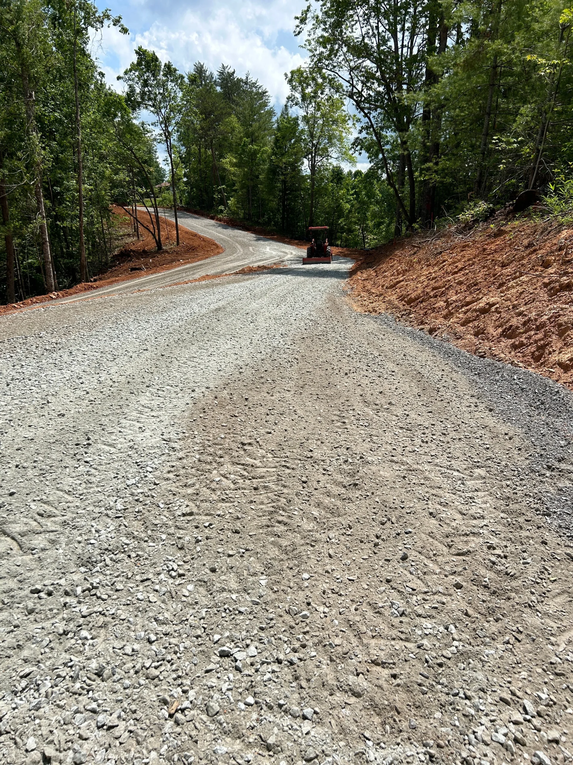 Gravel road winds through a wooded area, with a tractor working on the surface