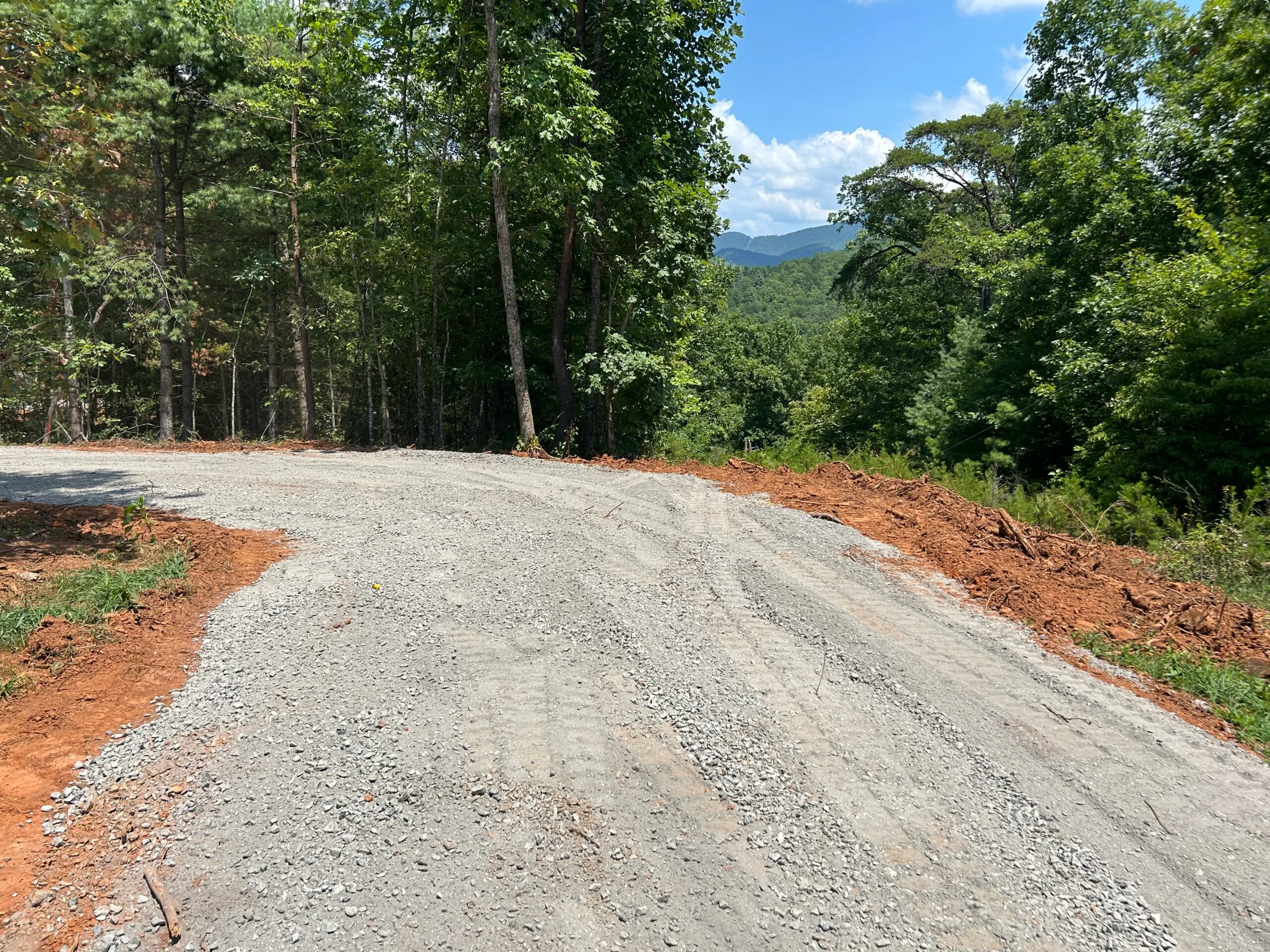 Gravel road winding through a forested area, with freshly cleared land on the sides and distant mountains visible under a clear blue sky.
