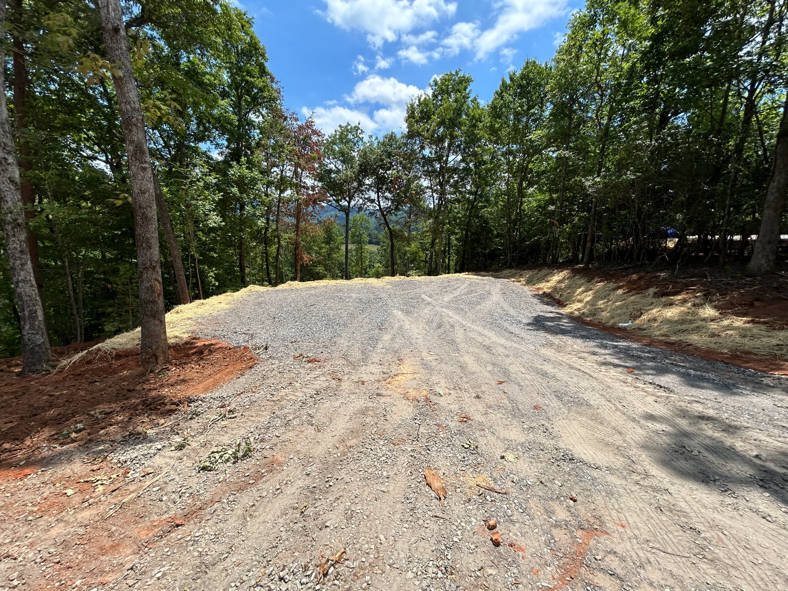 Cleared area with gravel foundation among trees, ready for construction, showcasing a scenic mountainous backdrop under a clear blue sky.