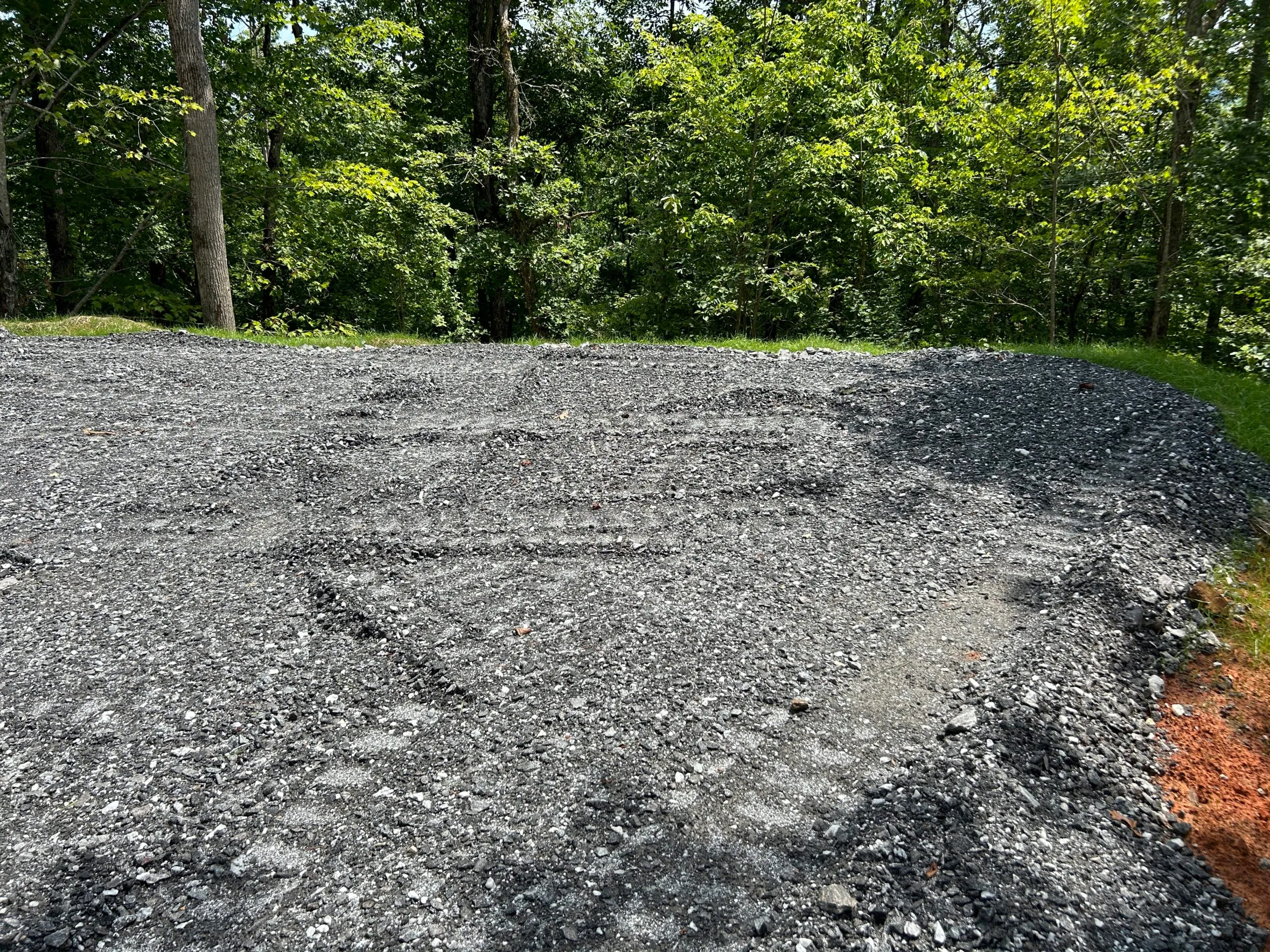 Cleared gravel area in a wooded setting, showing tire tracks and surrounding greenery