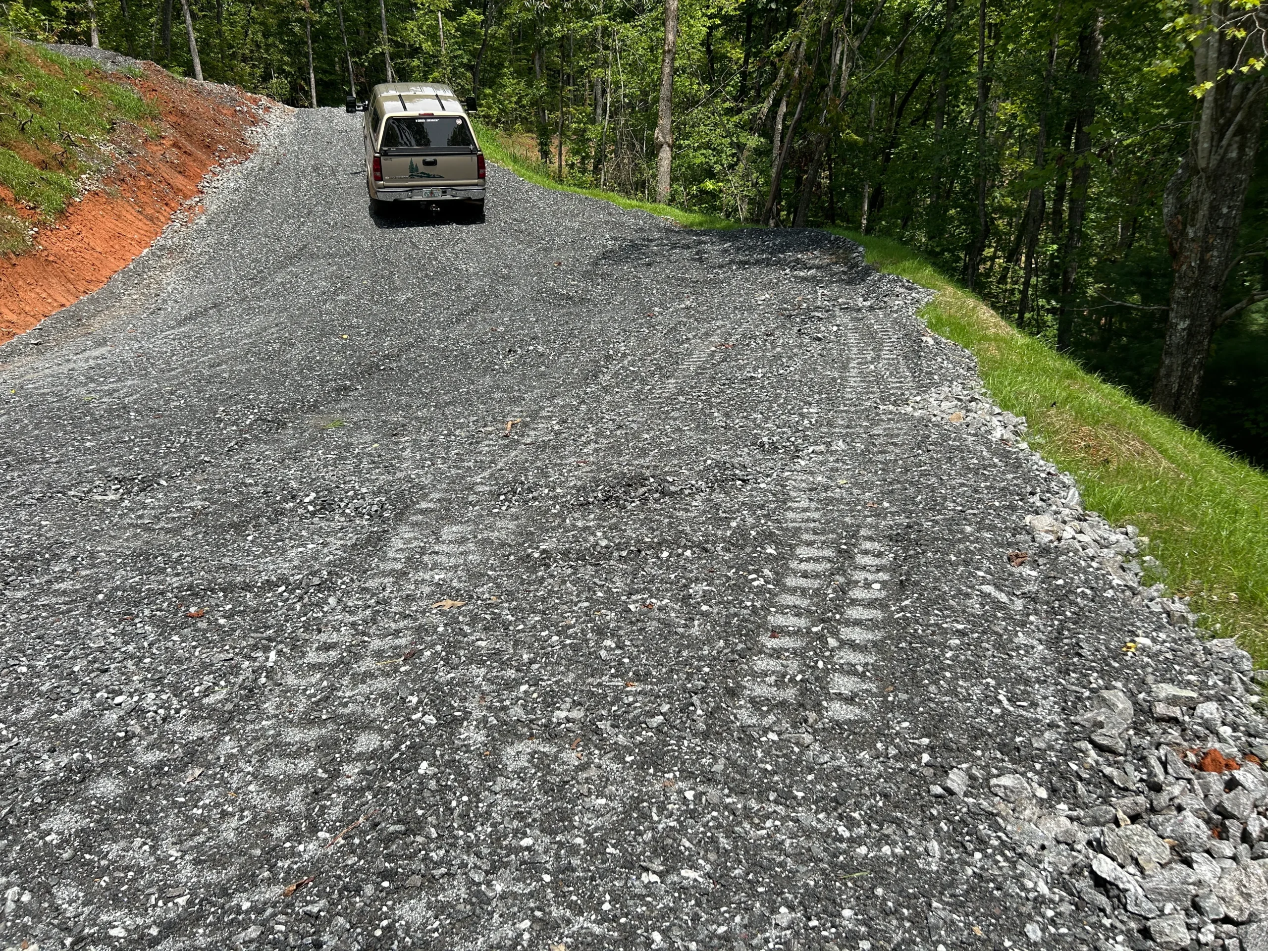 Gravel road curves uphill through a wooded area, with tire tracks visible. A vehicle is parked on the roadway